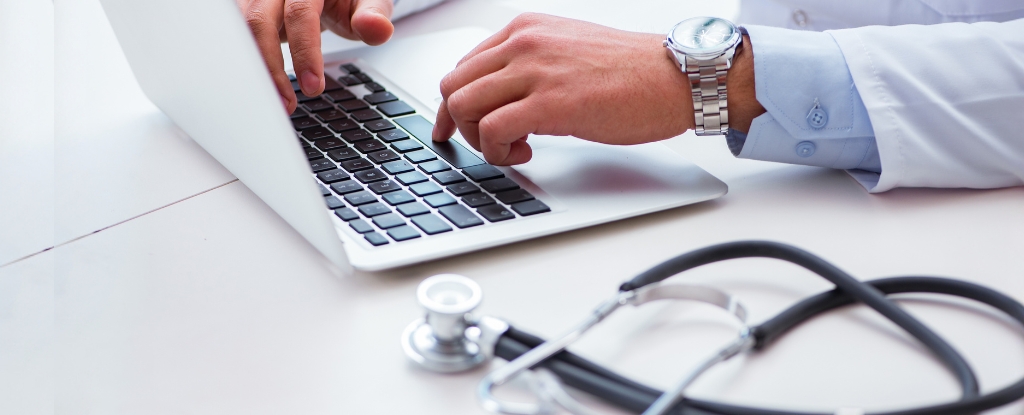 A laptop on a table next to a stethoscope. Hands are seen typing on the laptop, wearing a watch on the left wrist.