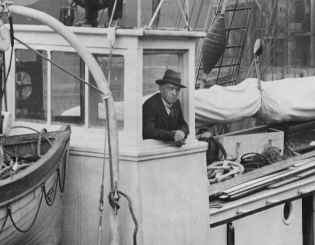 A black and white image of a man standing on a ship leaning out of a window