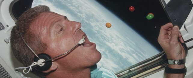 A man wearing a headset with an open mouth and lollies in air above it. Out the window the edge of Earth is visible from space