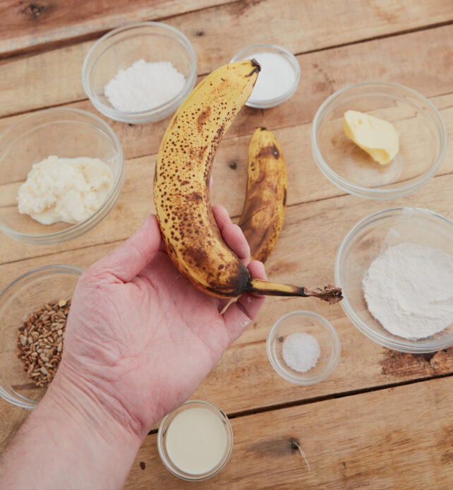 Person holding ripe banana in front of table with little glass bowls containing different baking ingrediants