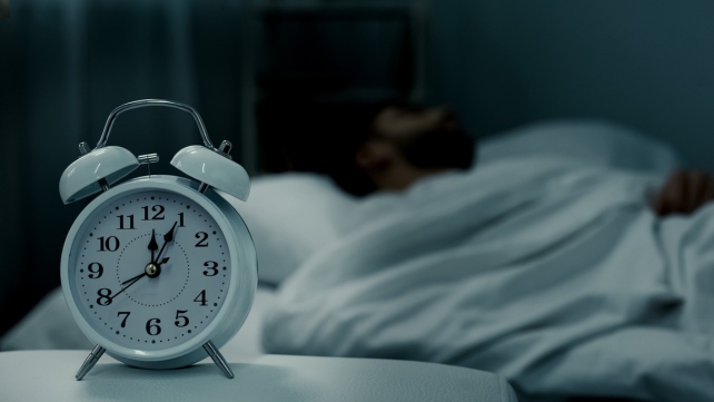 An alarm clock in the foreground with a blurred person in bed 