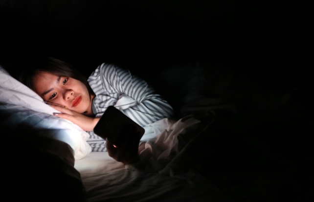 A brunette feminine person looking at a lit up phone in bed 