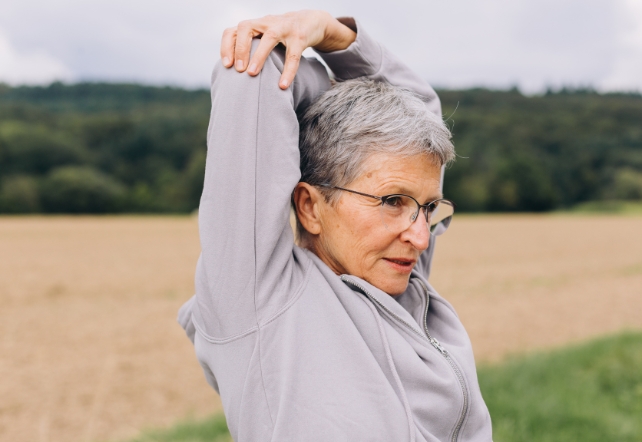 A senior woman with short grey hair and glasses, stretching with bent elbows behind head