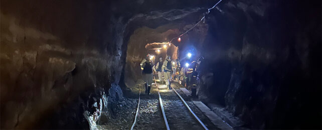 a team of people in protective gear and head torches walk away from the camera into the depths of a mine shaft. there are rail tracks at their feet.