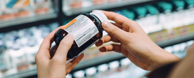 Hands holding pill bottle to investigate the label in front of pharmacy shelves