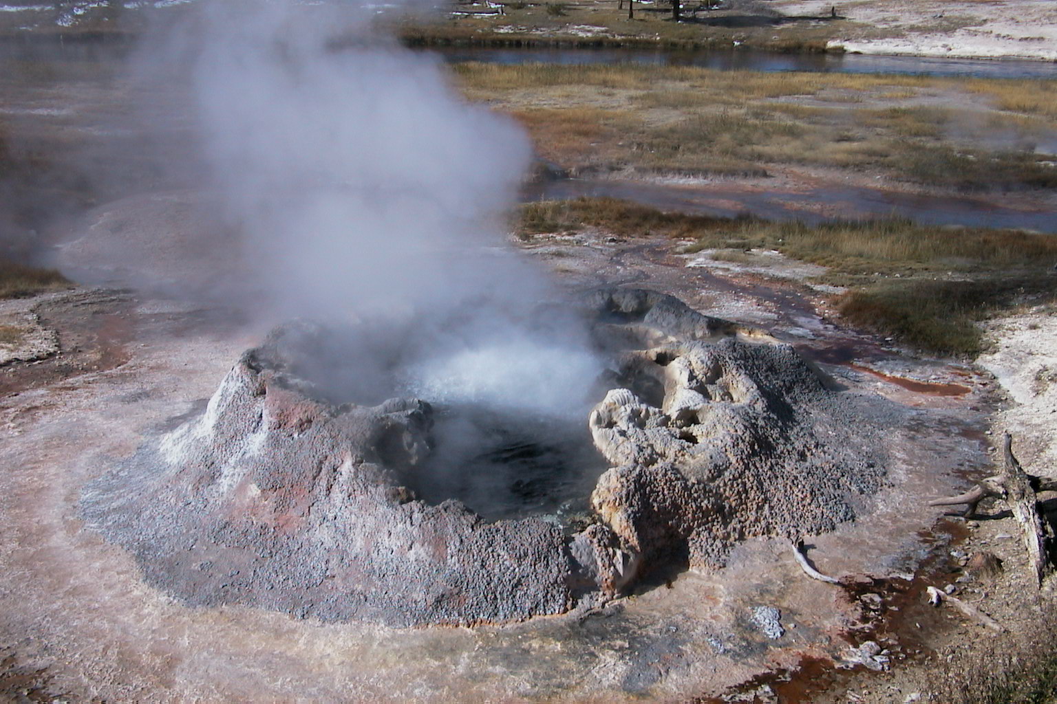 a hot spring with grey naturally-formed mineral walls around its rim, surrounded by flat rocky ground that appears slightly mossy. the hot spring has steam billowing from its center.