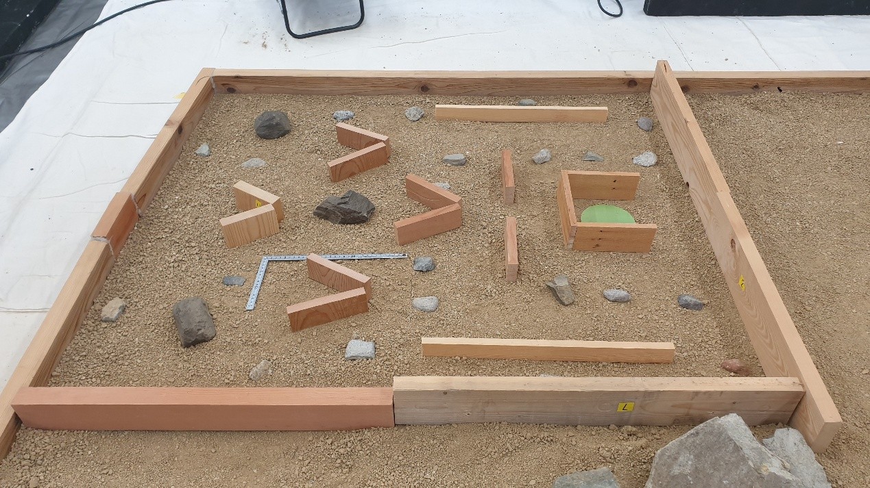 photo of a small 'arena' bounded by wood walls, lined with sand. various wood shapes, rocks and stones of different sizes are strewn across the sand. it appears to be in a lab.