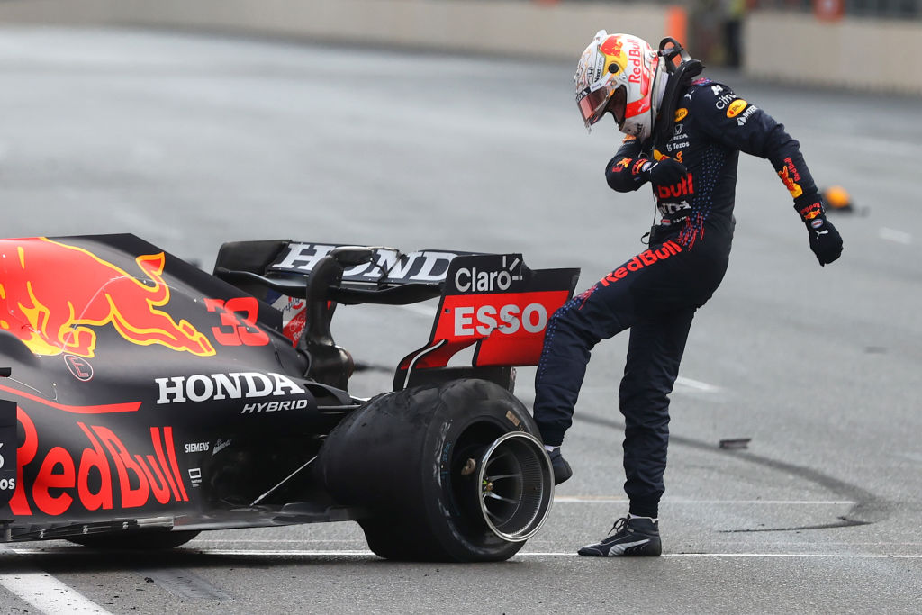 formula 1 racer max verstappen stands on the track kicking his race car's wheels. his uniform is black and so is the car, with 'honda' and 'redbull' logos printed across both.